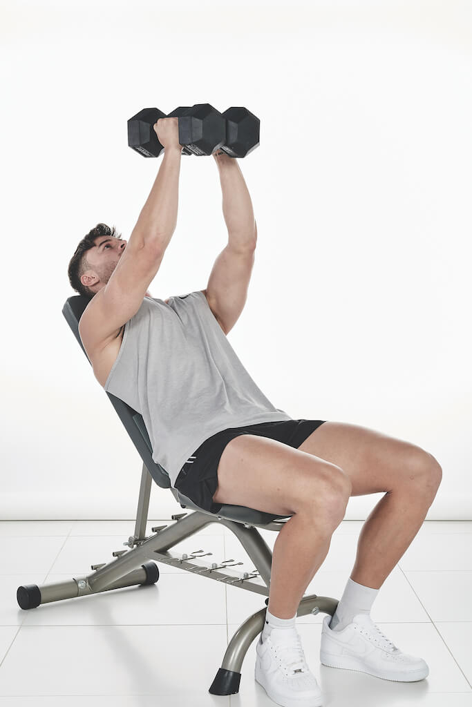 Man demonstrates how to do a dumbbell crush press; sitting on a bench at 45 degree incline, he holds a dumbbell in each hand; the dumbbells start on his chest, then he lifts them up above him before pressing them together and lowering with control 