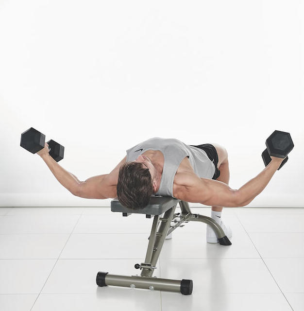 man demonstrates how to do a dumbbell flye: laying on a bench, he holds two dumbbells out either side of his body with bent arms; then he lifts them up to join in the middle above his body, before lowering to repeat