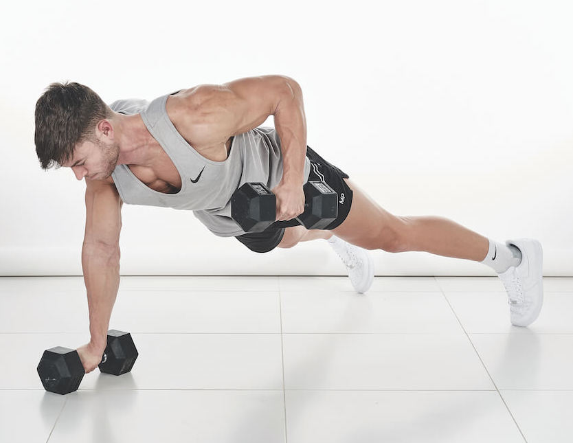 man demonstrates how to do a dumbbell press-up to renegade row: in a plank position, with two dumbbells on the floor directly below his shoulders, he lowers his body to the ground before rising again and lifting the left dumbbell towards him; he lowers, then repeats on the other side