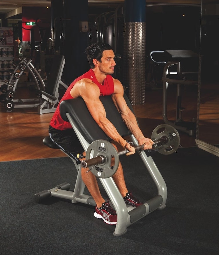 man in red vest and shorts performing the first stage of an EZ bar preacher curl in the gym