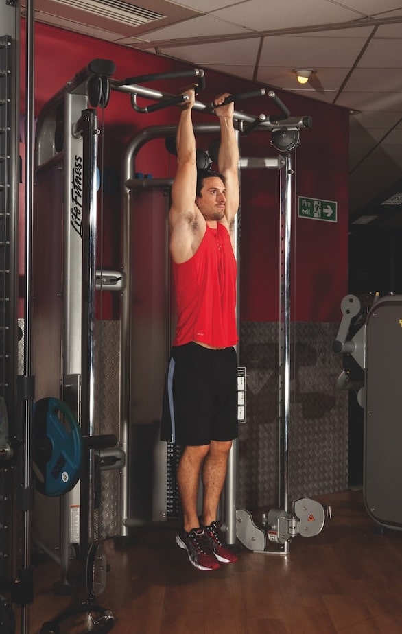 man in red vest and shorts performing the first stage of a hanging leg raise in the gym
