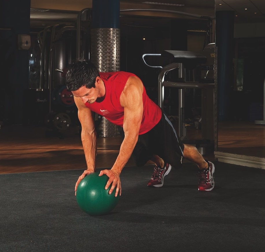 man in red vest and shorts performing the first stage of a medicine ball press-up in the gym