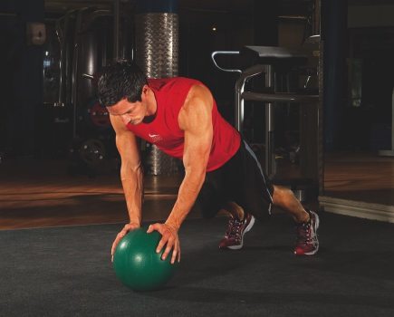 man in red vest and shorts performing the first stage of a medicine ball press-up in the gym