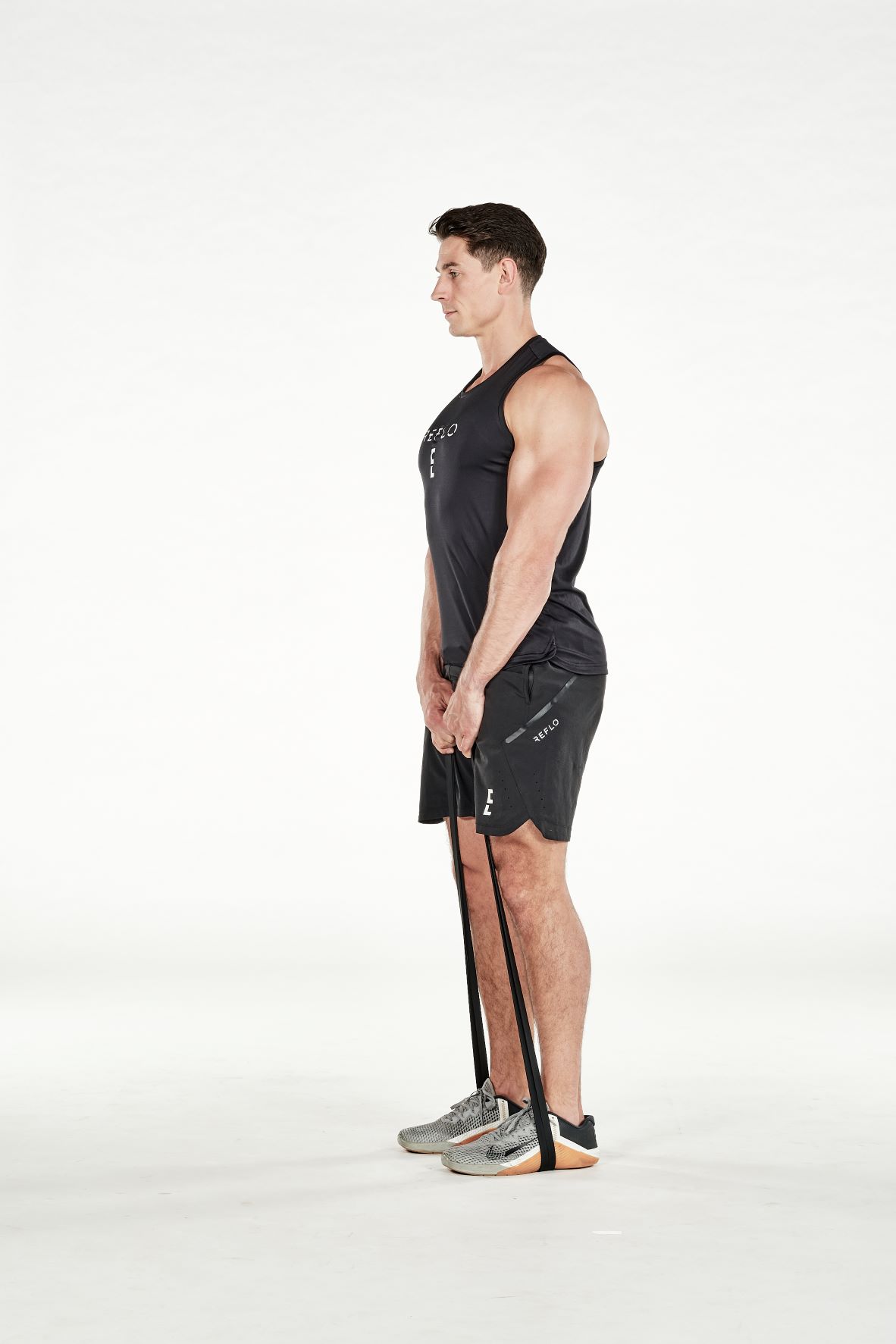 man demonstrating step two of band deadlift; he is standing up straight, holding a band that is wrapped around his feet; he wears a black fitness vest, black shorts and trainers