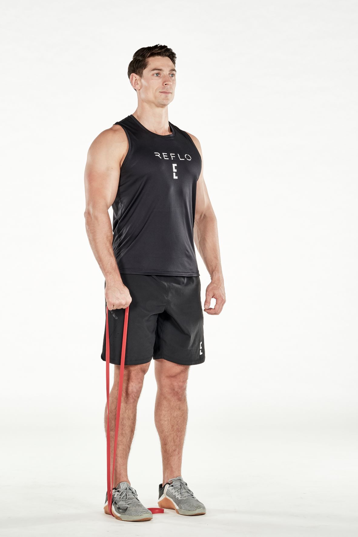 man demonstrating step one of resistance band front raise; standing up straight, he is holding a resistance band that is wrapped under one foot in one hand; he wears a black fitness vest, black shorts and trainers