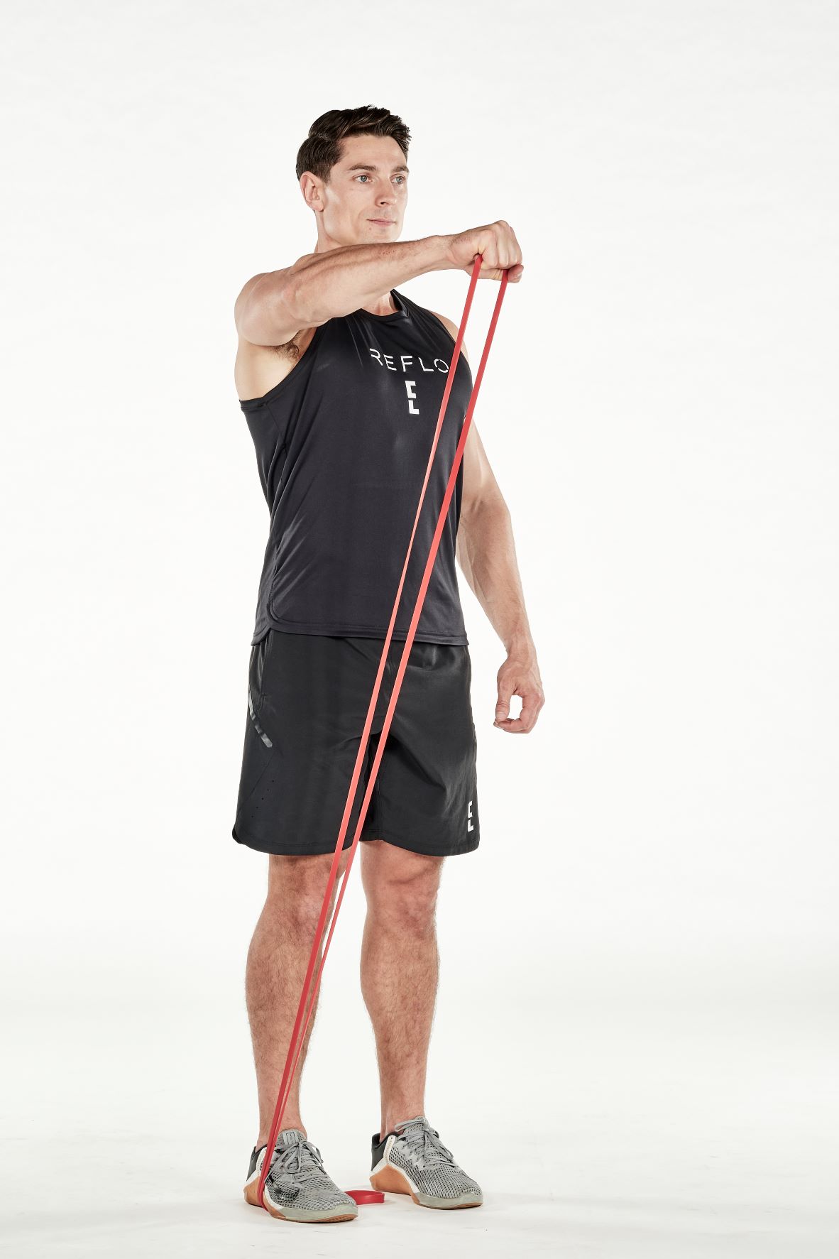 man demonstrating step two of resistance band front raise; standing up straight, he is holding a band that is wrapped under one foot in one hand; keeping the arm straight, he lifts and extends his arm up until it is level with his shoulder; he wears a black fitness vest, black shorts and trainers