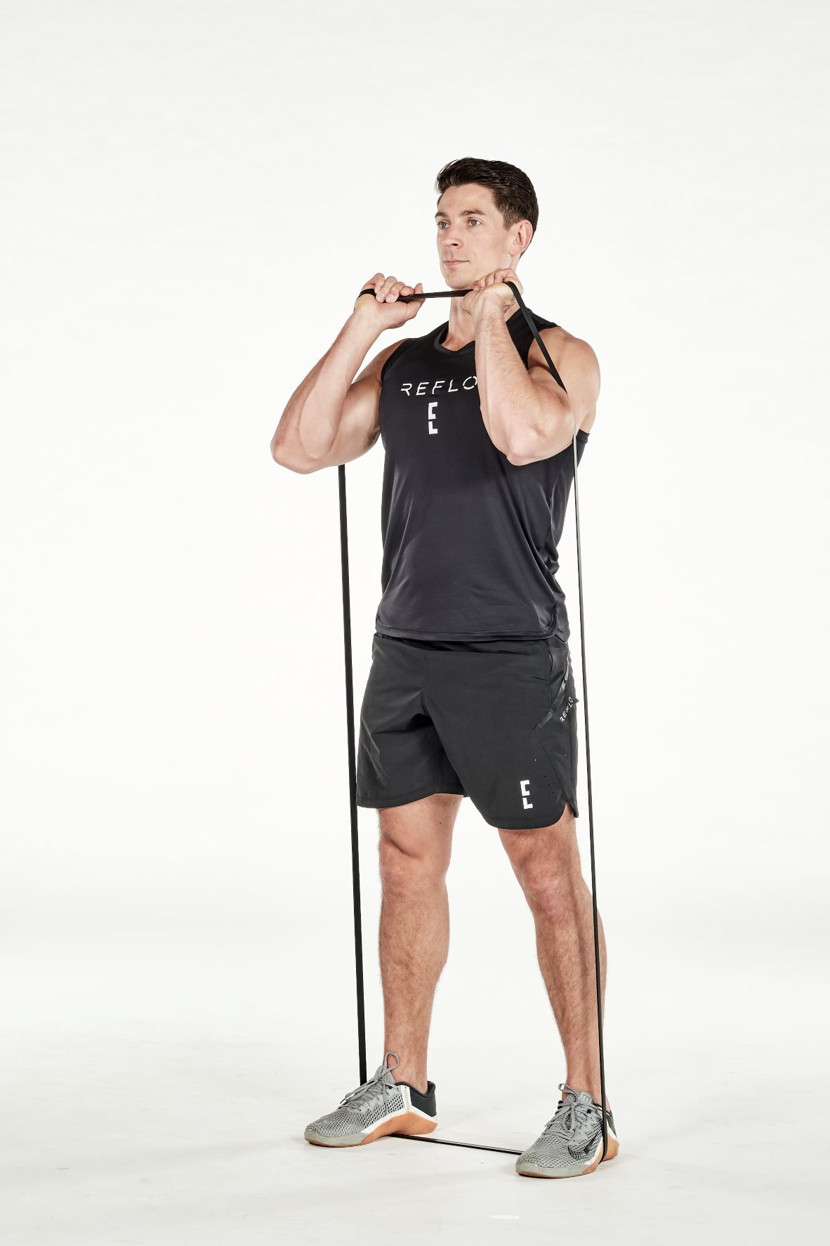 man demonstrating step one of resistance band front squat; standing up straight, he is holding a resistance band that is wrapped under both feet in two hands, holding it level with his shoulders; he wears a black fitness vest, black shorts and trainers