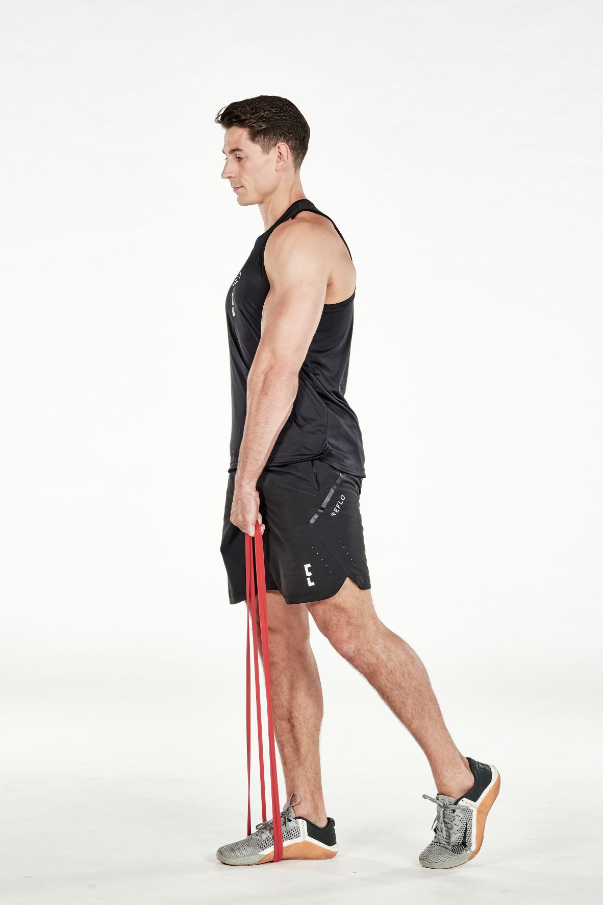man demonstrating step two of resistance band single-leg romanian deadlift; he stands straight, holding a band which is being held under his foot; his other leg is relaxed and positioned behind him; he wears a black fitness vest, black shorts and trainers