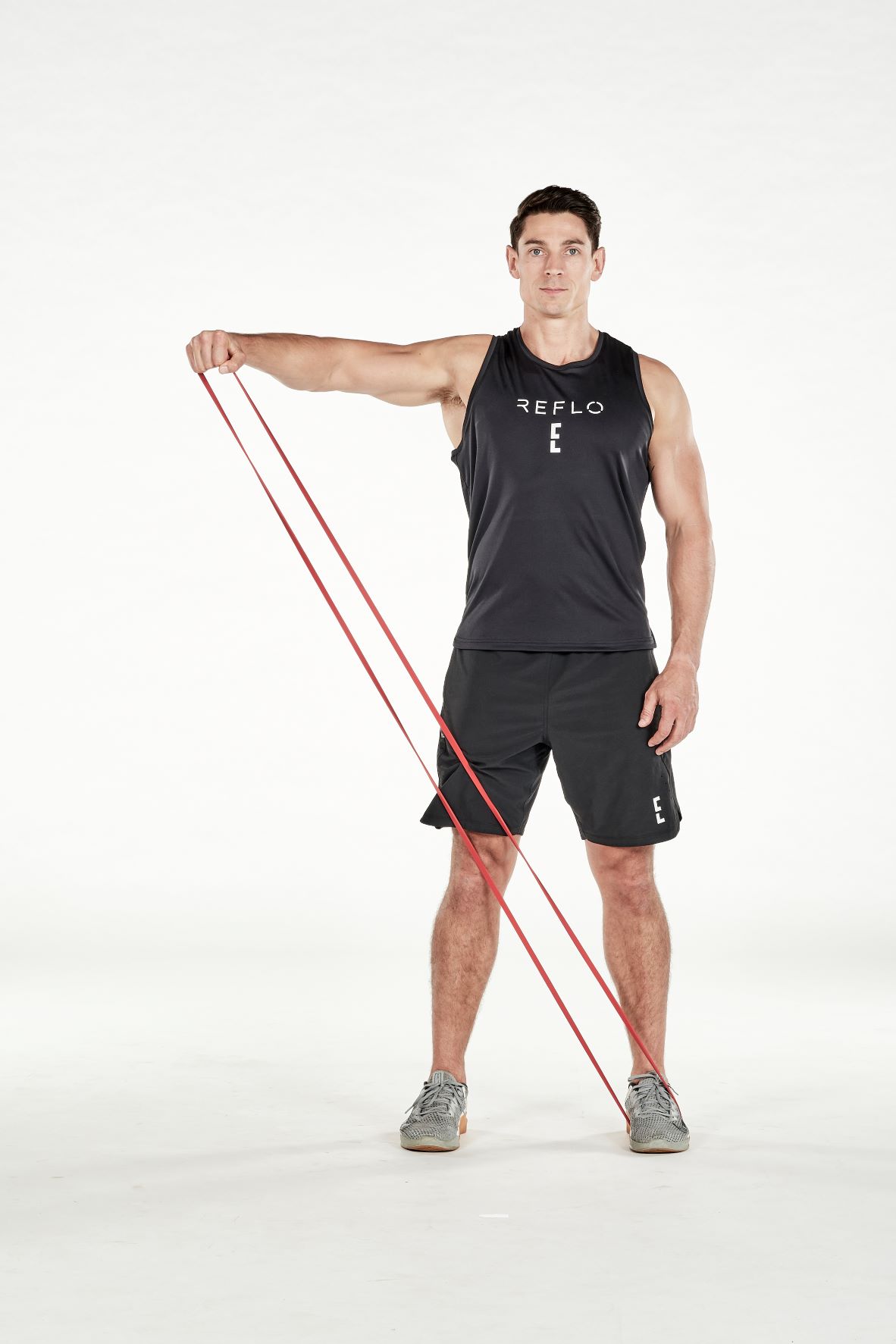 man demonstrating step two of resistance band lateral raise; standing up straight, a resistance band is held under one foot; holding the resistance band with the opposite hand, he extends his arm straight out to the side until it reaches shoulder height; he wears a black fitness vest, black shorts and trainers