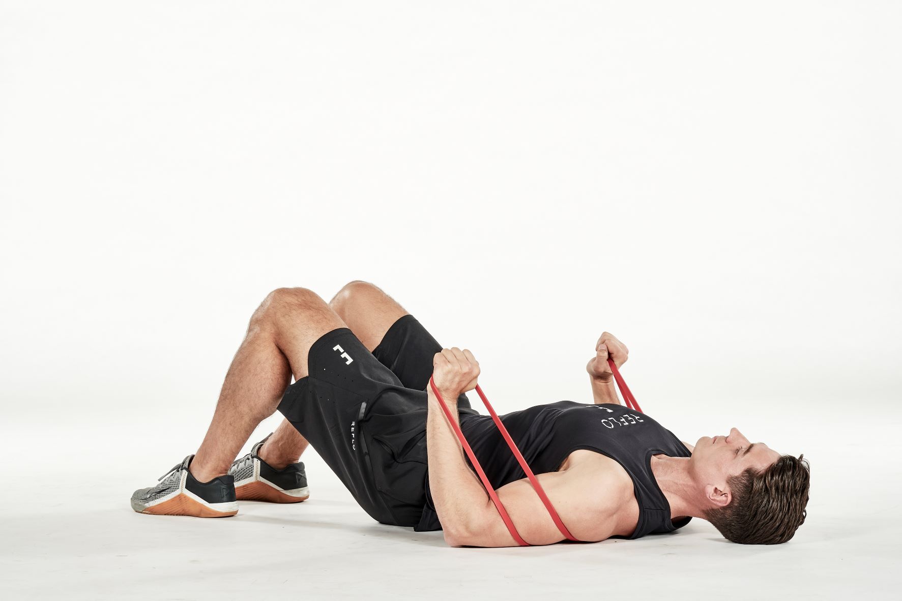 man demonstrating step one of lying chest press; laying on his back with bent knees, the resistance band is passed under his back; his arms are bent as each hand holds one end of the resistance band; he wears a black fitness vest, black shorts and trainers