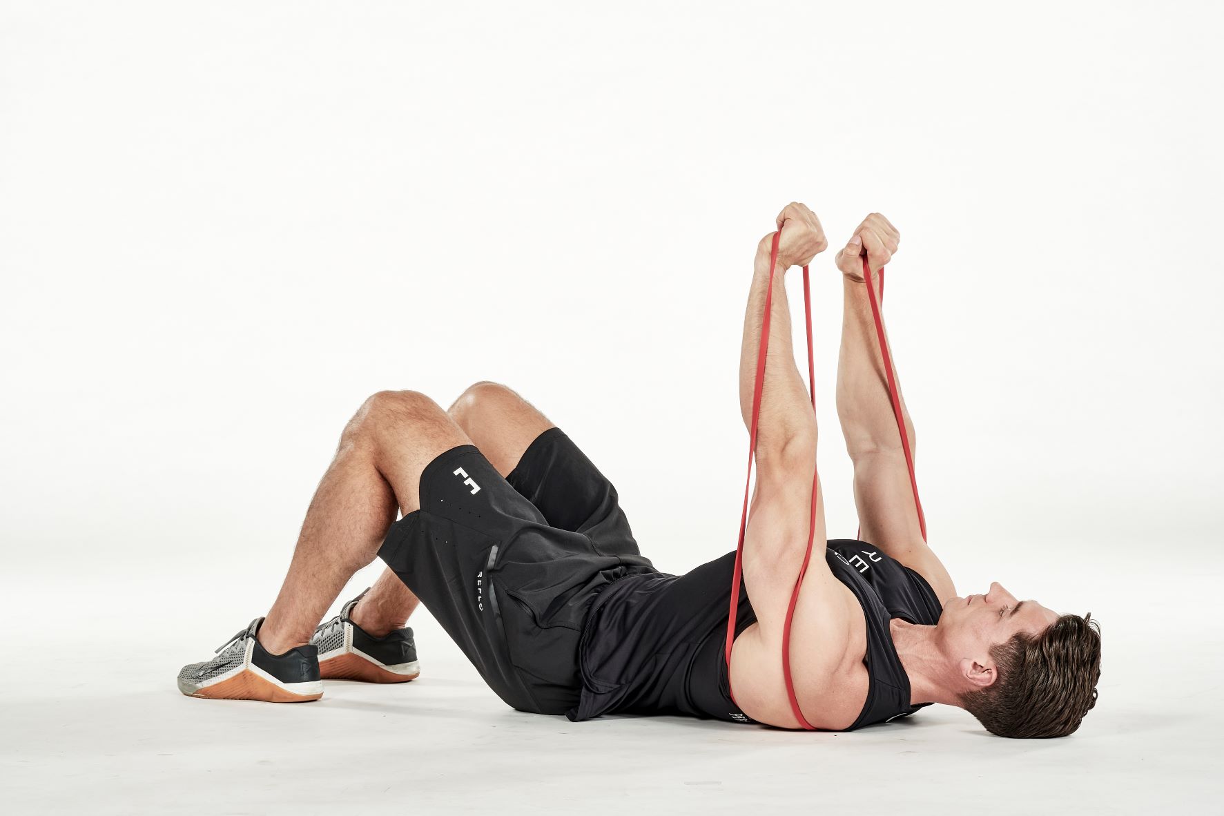 man demonstrating step two of lying chest press; laying on his back with bent knees, the band is passed under his back; holding one end of the resistance band in each hand, he extends his arms up towards the ceiling; he wears a black fitness vest, black shorts and trainers