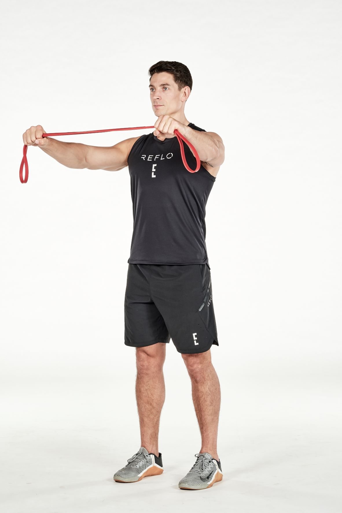 man demonstrating step one ofreverse fly; standing up straight, he holds a resistance band out in front of him in both hands, shoulder width apart; he wears a black fitness vest, black shorts and trainers