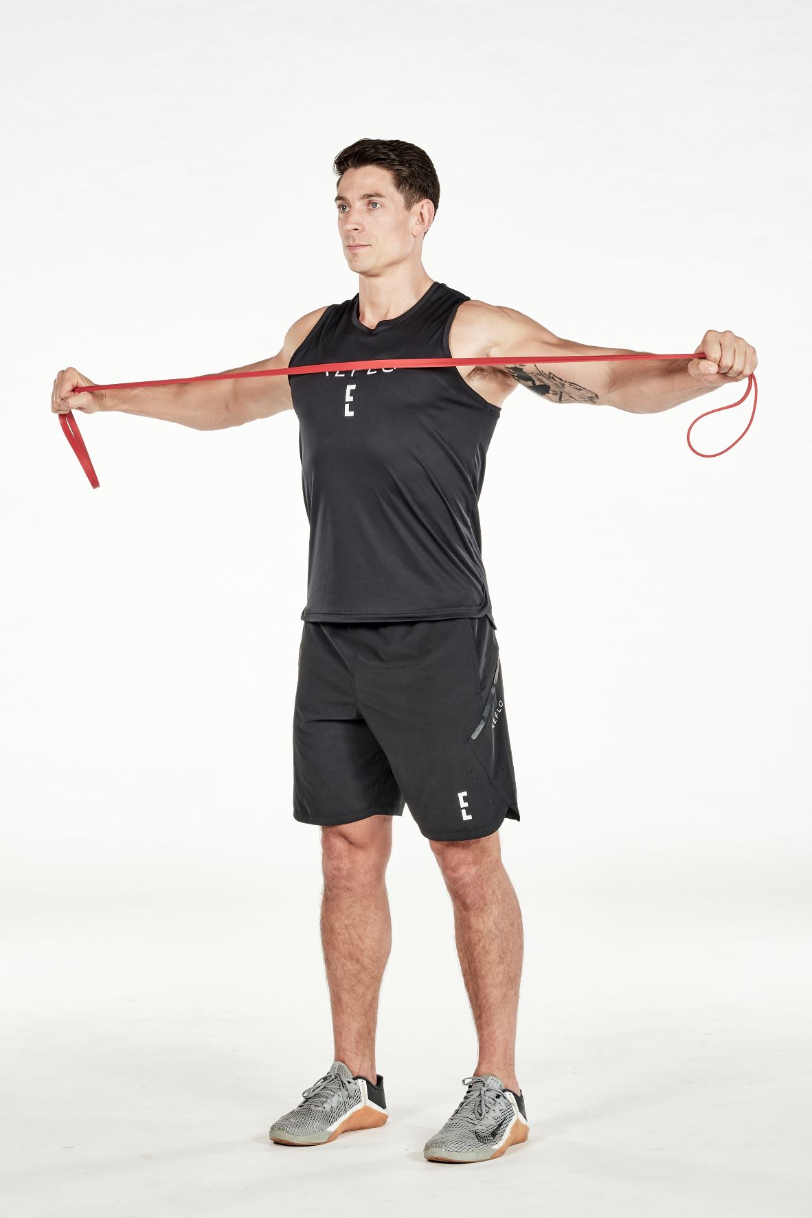 man demonstrating step two of reverse fly; standing up straight, he holds a band in both hands, he stretches his arms outwards as far as possible; he wears a black fitness vest, black shorts and trainers