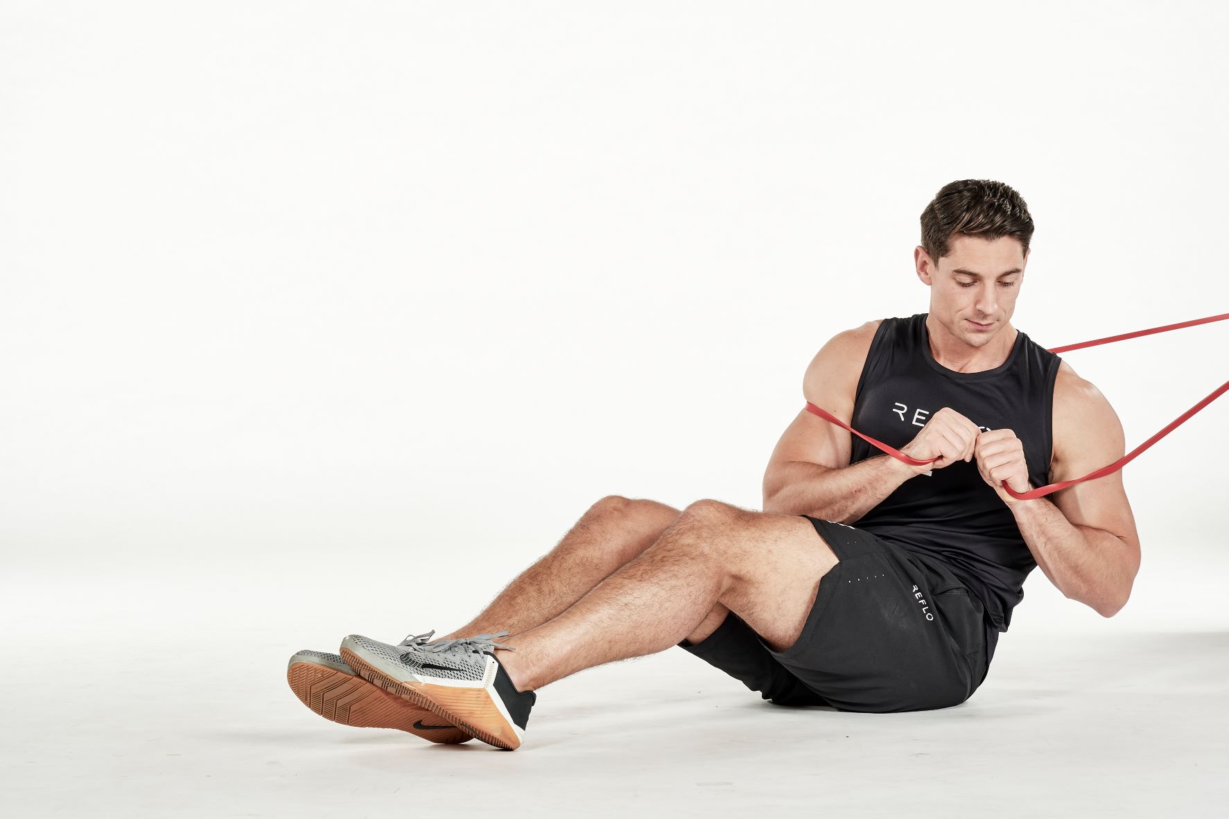 man demonstrating step two of russian twist; sitting down with knees bent, he holds a secured band in his hands in front of his chest; he turns his body to one side; he wears a black fitness vest, black shorts and trainers