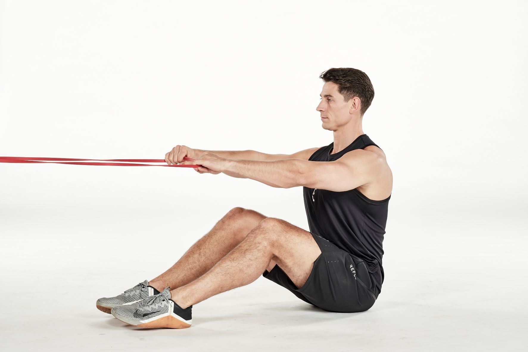man demonstrating step one of seated face pull; seated with knees bent, he holds a secured resistance band; his arms are straight, outstretched in front of him; he wears a black fitness vest, black shorts and trainers
