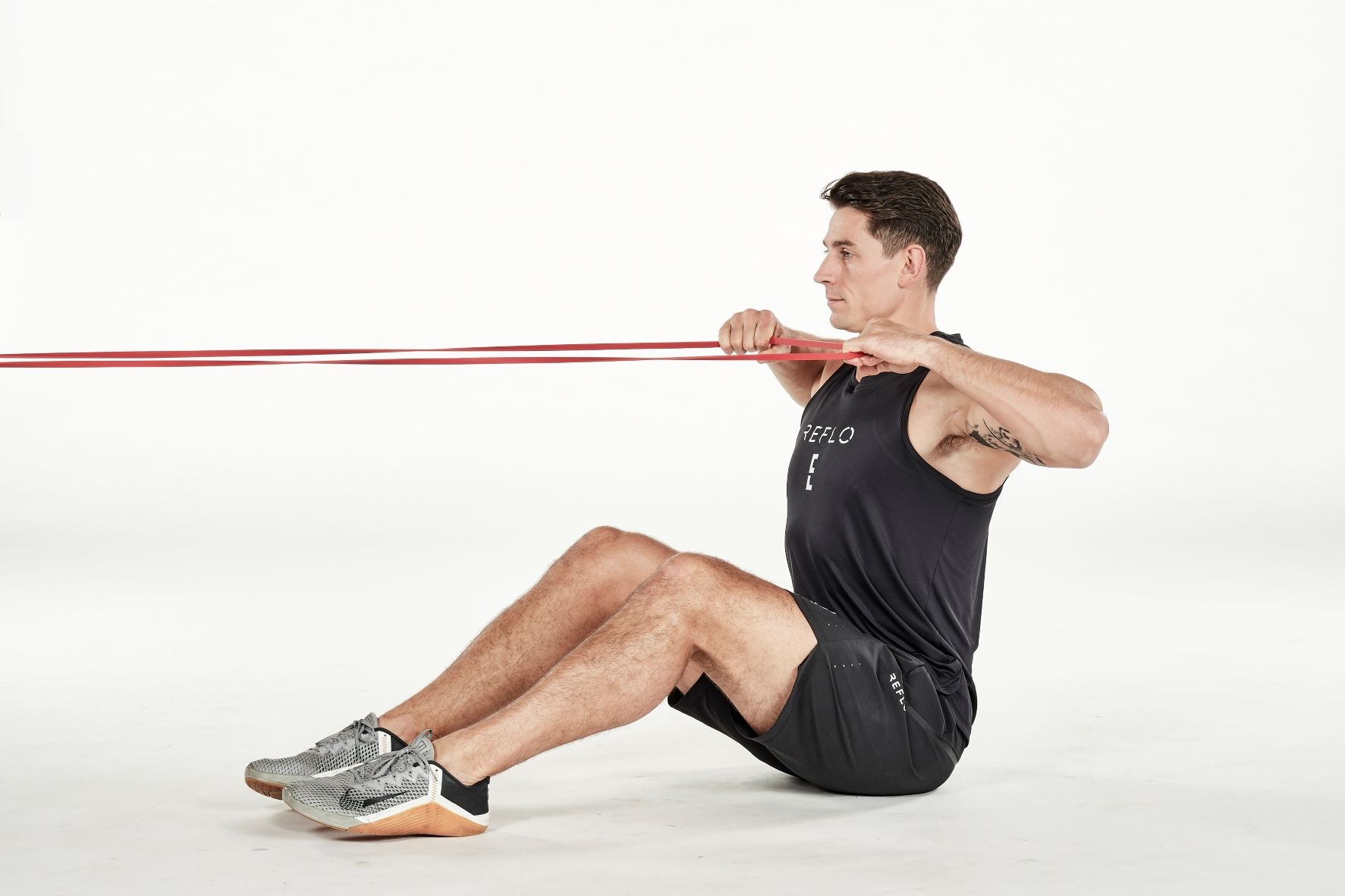 man demonstrating step two of seated face pull; seated with knees bent, he holds a secured band; his arms are bent as he pull the band towards his chest; he wears a black fitness vest, black shorts and trainers
