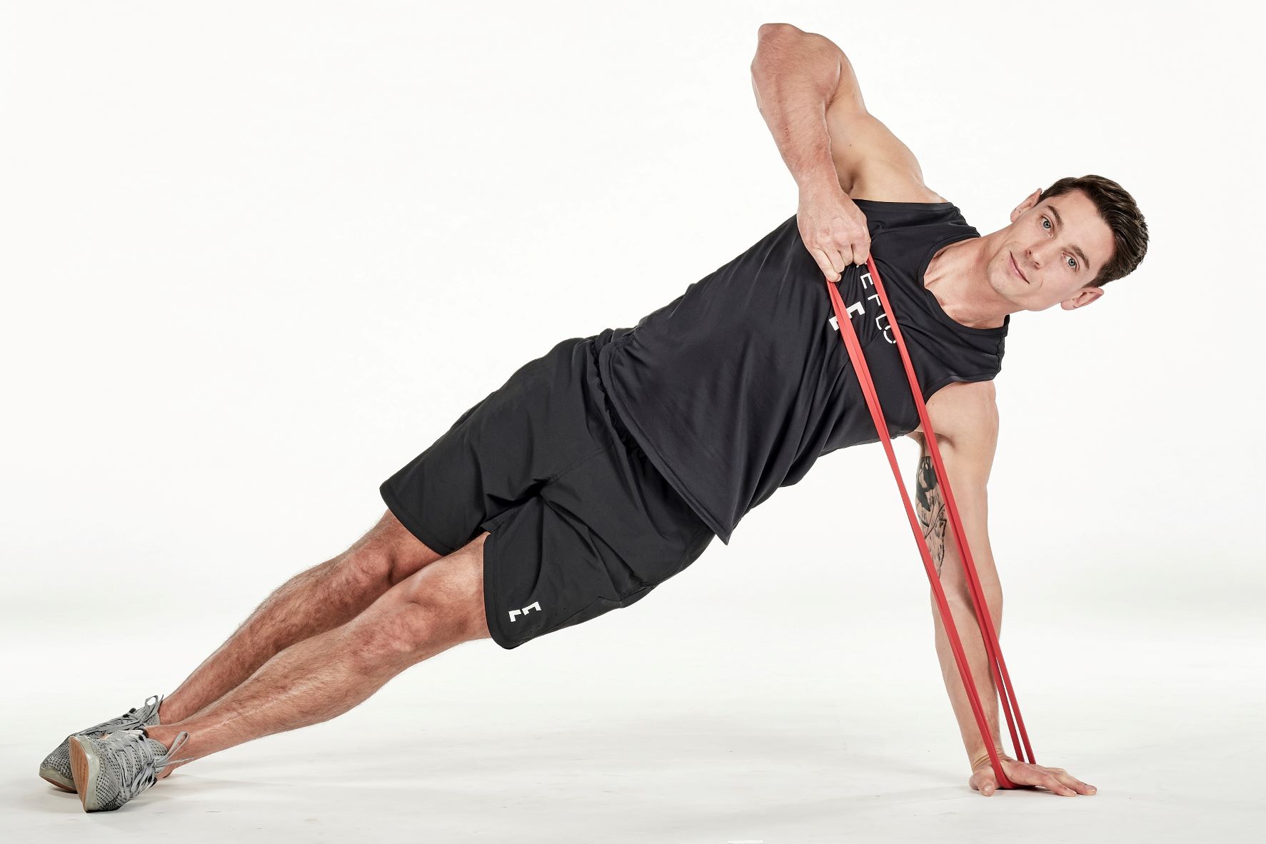 man demonstrating step two of side plank high pull; in a side plank position, his top hand holds a band that is secured under his bottom hand on the floor; his top arm is bent as he pulls the band upwards; he wears a black fitness vest, black shorts and trainers