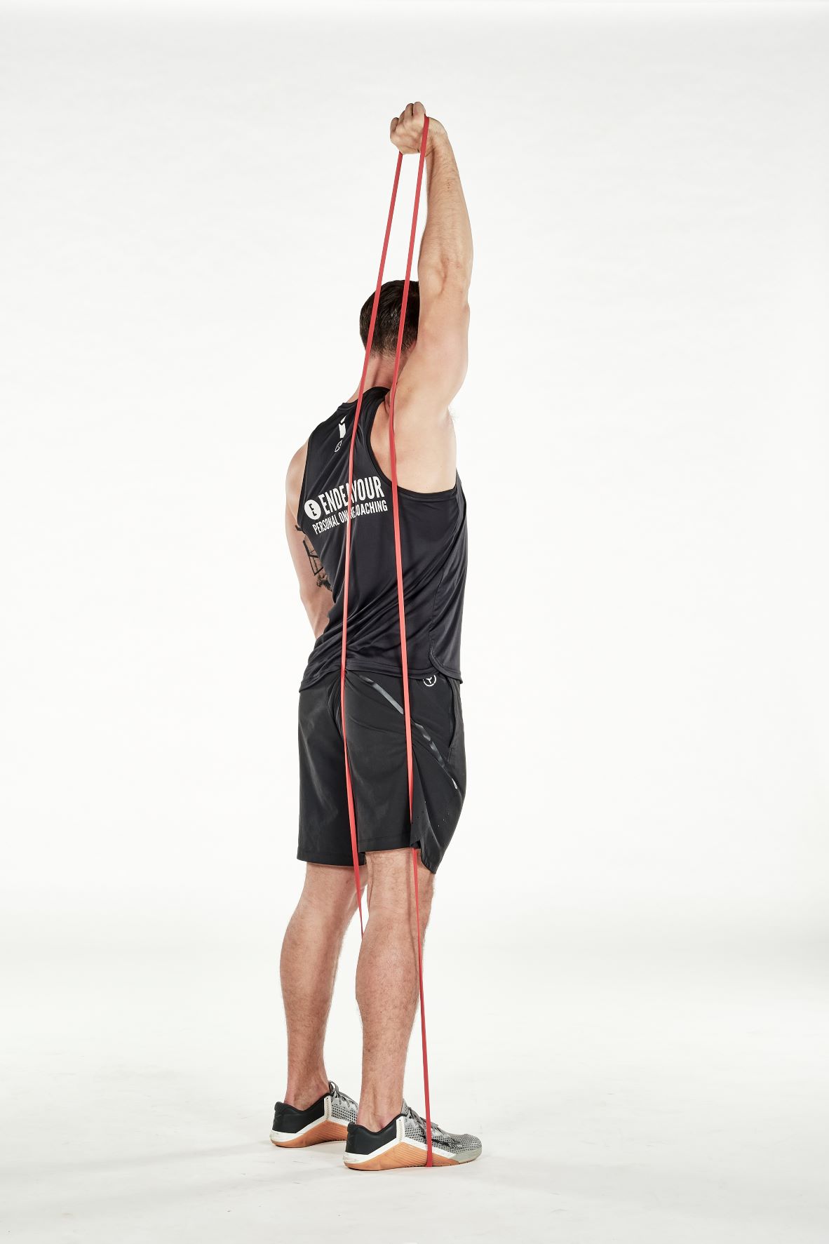 man demonstrating step two of single arm triceps extension; standing tall, his arm reaches behind his head to hold a band that's held under one foot; his arm straightens as he pulls the band and reaches up; he wears a black fitness vest, black shorts and trainers