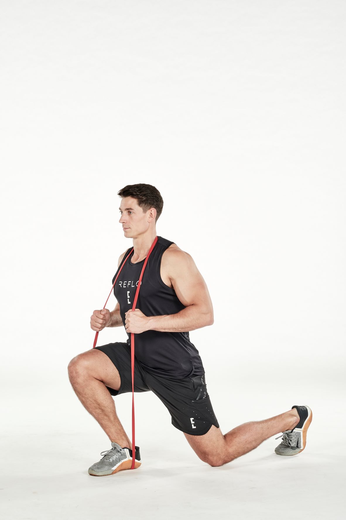man demonstrating step two of split squat; bending down into a lunge position, he holds a band under his front foot and around the back of his neck; his hands support the band in the middle; his arm is bent; he wears a black fitness vest, black shorts and trainers