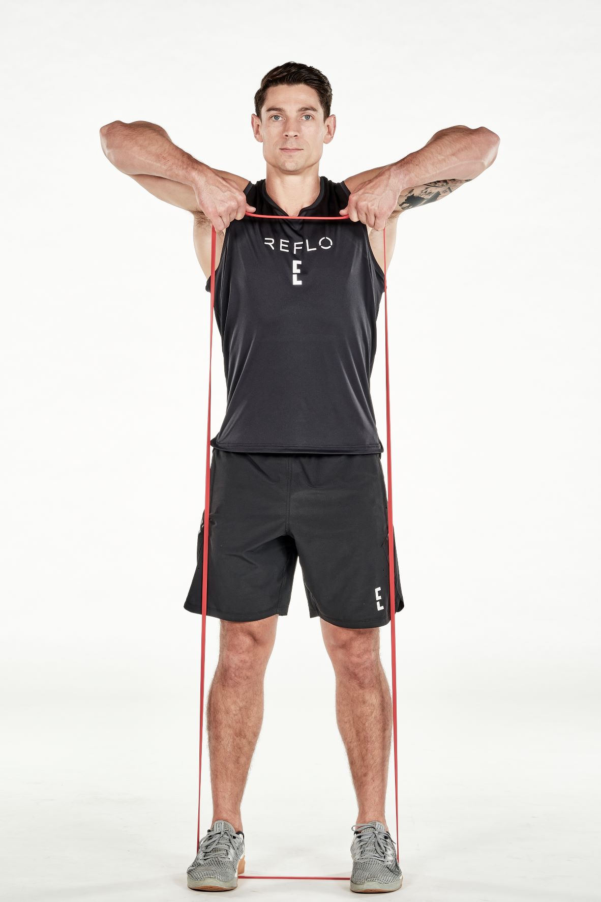 man demonstrating step two of upright row; he stands upright, both hands holding a band that is secured under his feet; he pulls up on the band until level with his shoulders; he wears a black fitness vest, black shorts and trainers