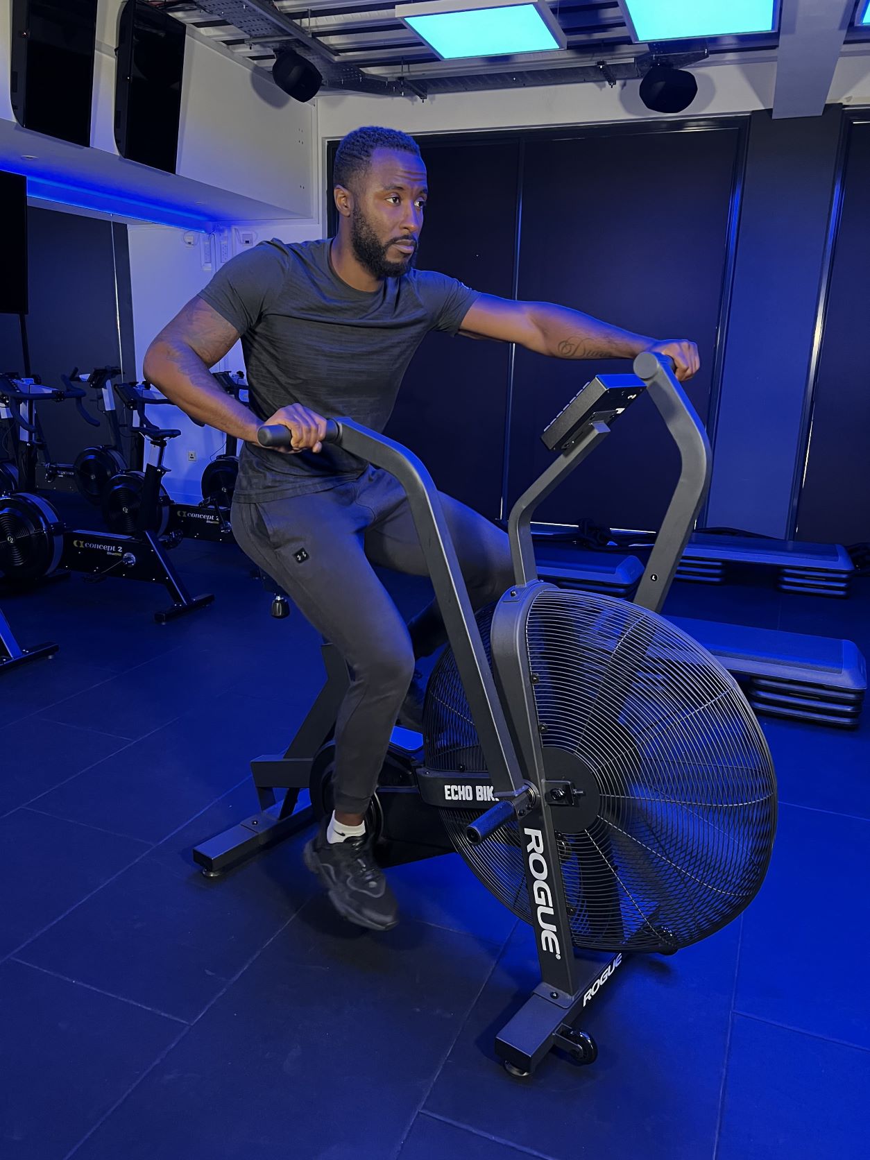 man demonstrates how to use an air bike; he sits on the bike with his legs pedalling and arms pushing and pulling the two handles in opposite directions