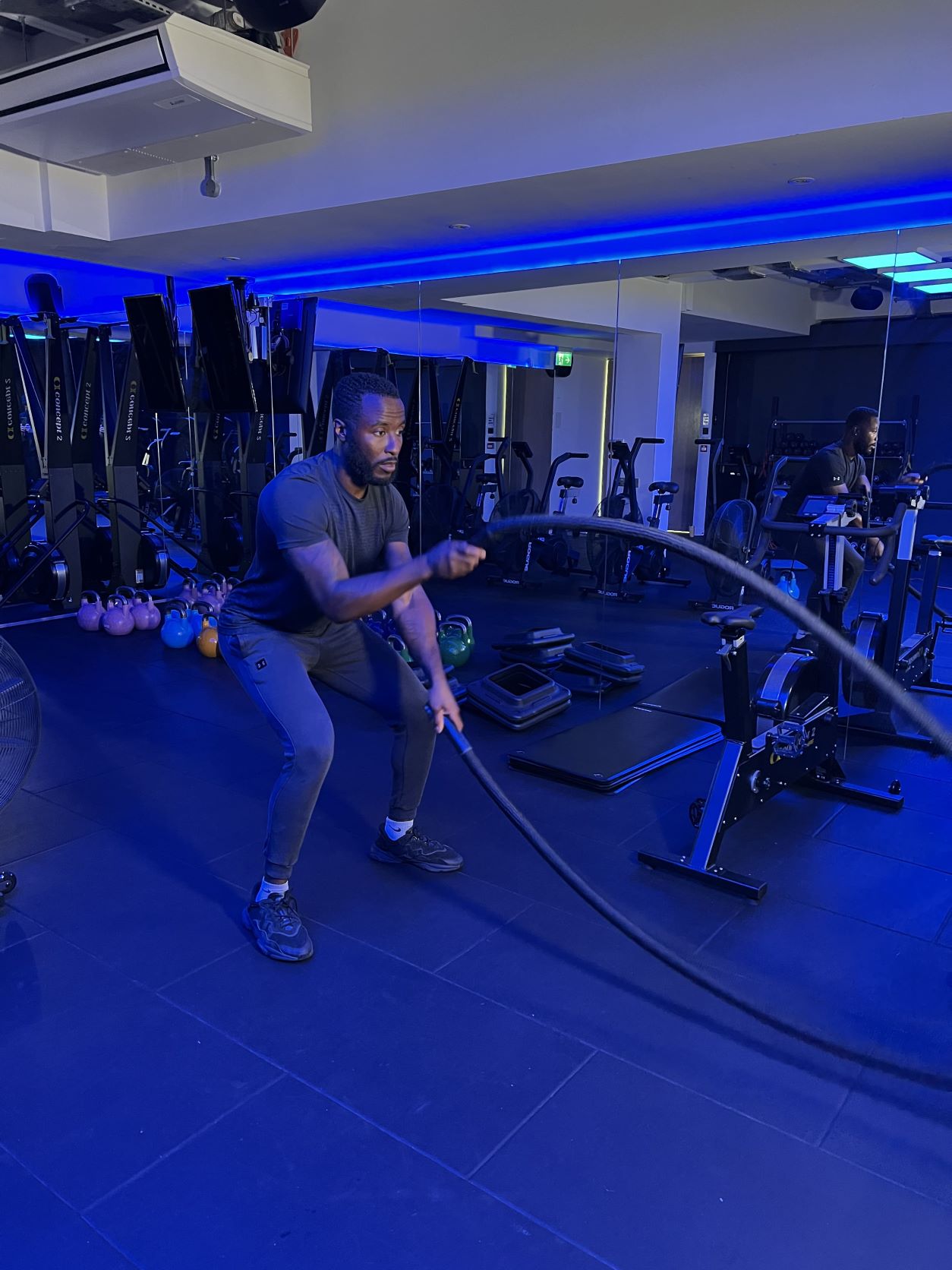 man demonstrates how to use battle ropes at the gym; bent over, he holds a heavy rope in each hand and alternately raises and lowers each one