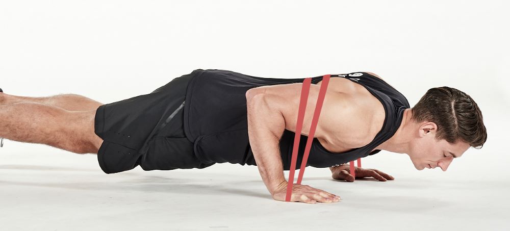 man demonstrating step two of press up; with the band passing over his back to be held by each hand on the floor, he bends his arms to lower his body towards the floor; he wears a black fitness vest, black shorts and trainers