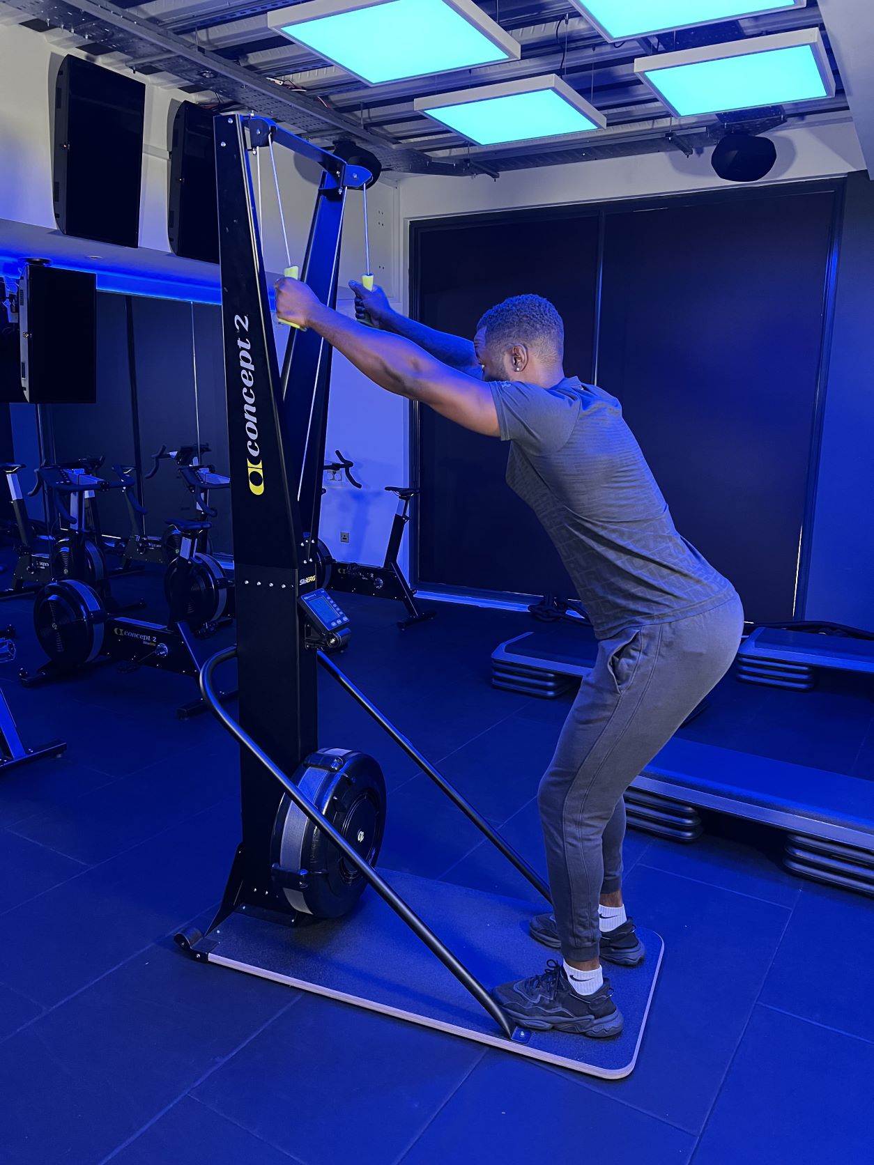 man demonstrating how to use a skierg machine at the gym; he holds the cables in both hands above his head