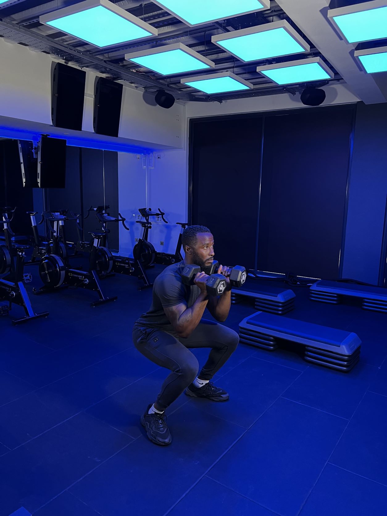 man demonstrates how to do a squat press at the gym; step 2, he squats low while holding a dumbbell in each hand at shoulder height