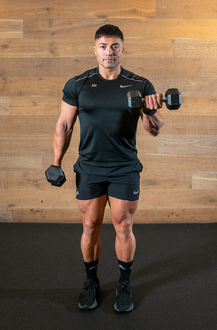Part of the best full-body dumbbell workout: man demonstrated dumbbell curls; standing upright, he holds a dumbbell in each hand and alternately lifts each one towards his chest