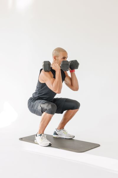 front squat demonstration: man holds two dumbbells at shoulder height, he bends his knees to squat