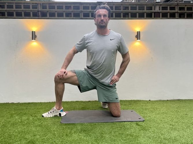 man demonstrates step one of the adductor dip stretching exercise. He kneels on one knee, with the other thigh turned out.