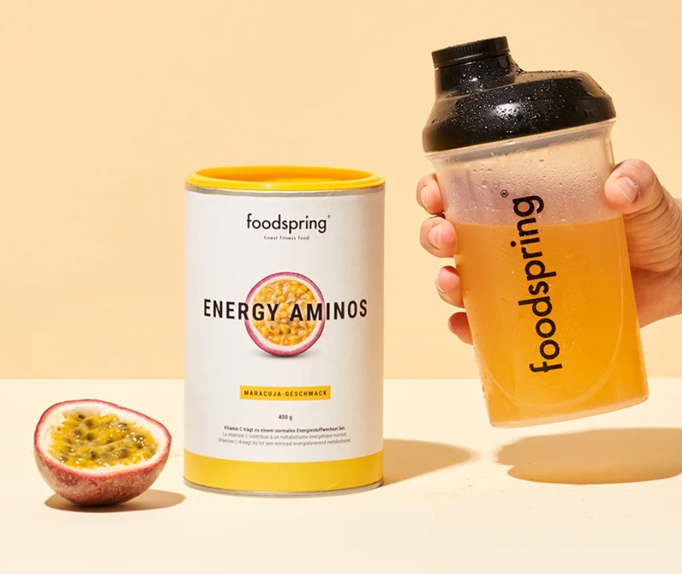 Product shot of a tub of Foodspring Energy Amino powder, a hand with a drink bottle and half a passionfruit