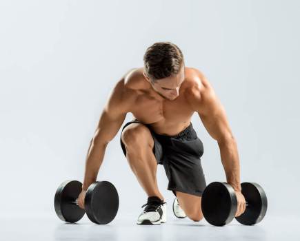 Topless man kneeling on the floor with a pair of dumbbells