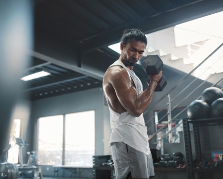 man using dumbbells in gym workout, wearing white tank top and grey shorts