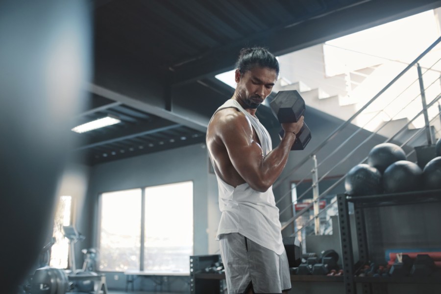 man using dumbbells in gym workout, wearing white tank top and grey shorts