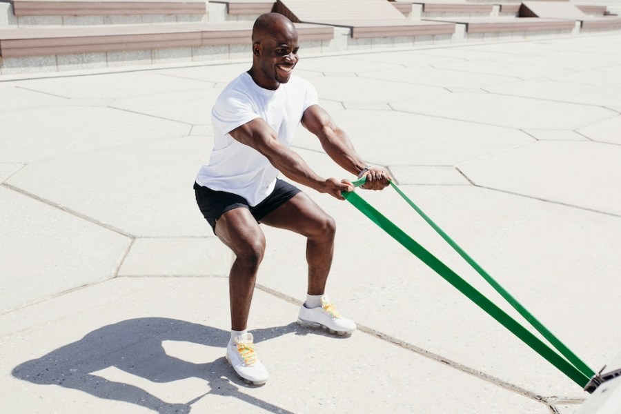 Afro American man in white t-shirt and black shorts doing exercise on his hands squatting with elastic fitness tape outside