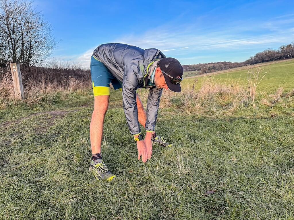 Running coach Simon James performing hamstrings stretch in a field after a trail run