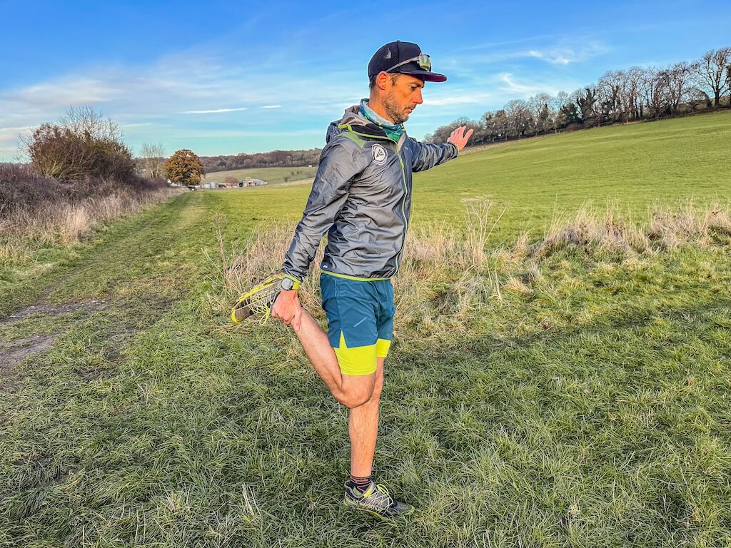 Running coach Simon James performing quads stretch in a field after a trail run