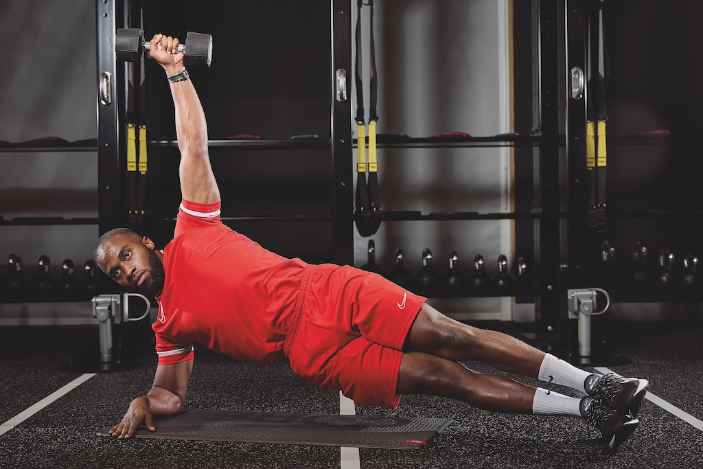 man in red top and red shorts doing a side plank with dumbbell triceps extension