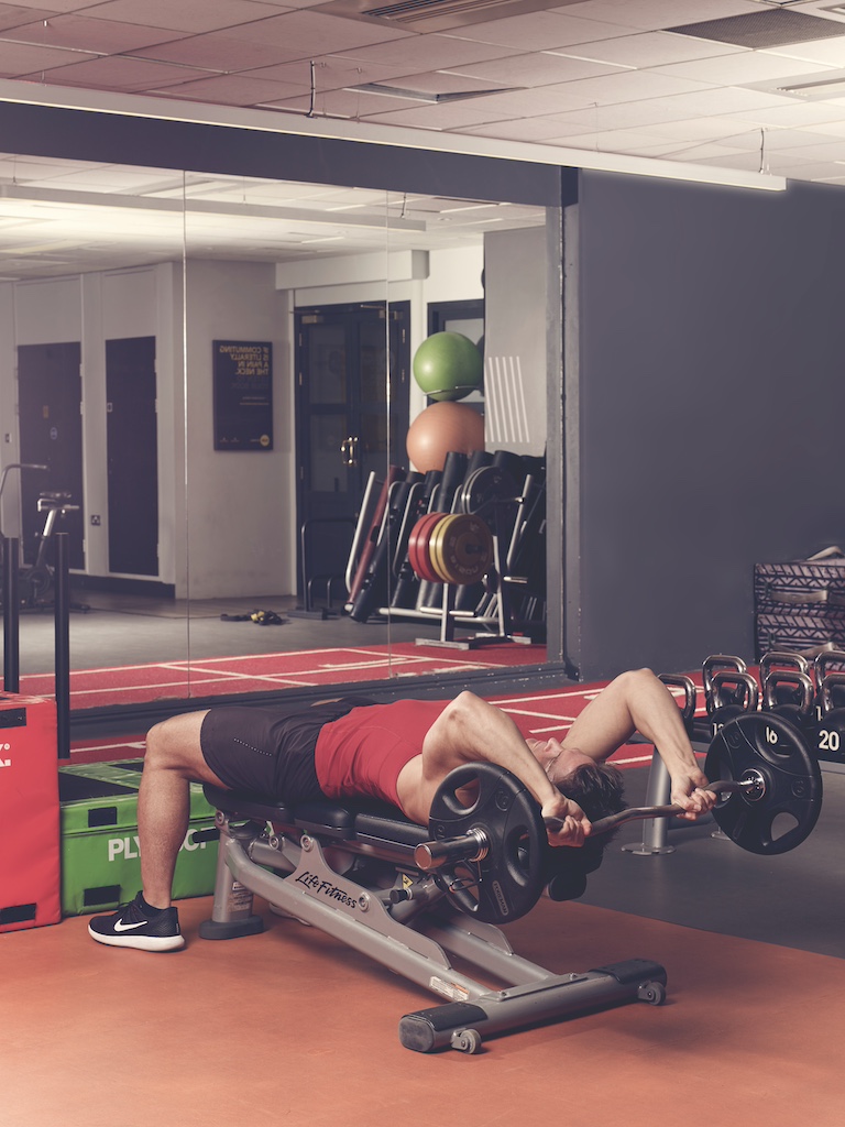 man in red vest and black shorts performing EZ-bar triceps extension in the gym