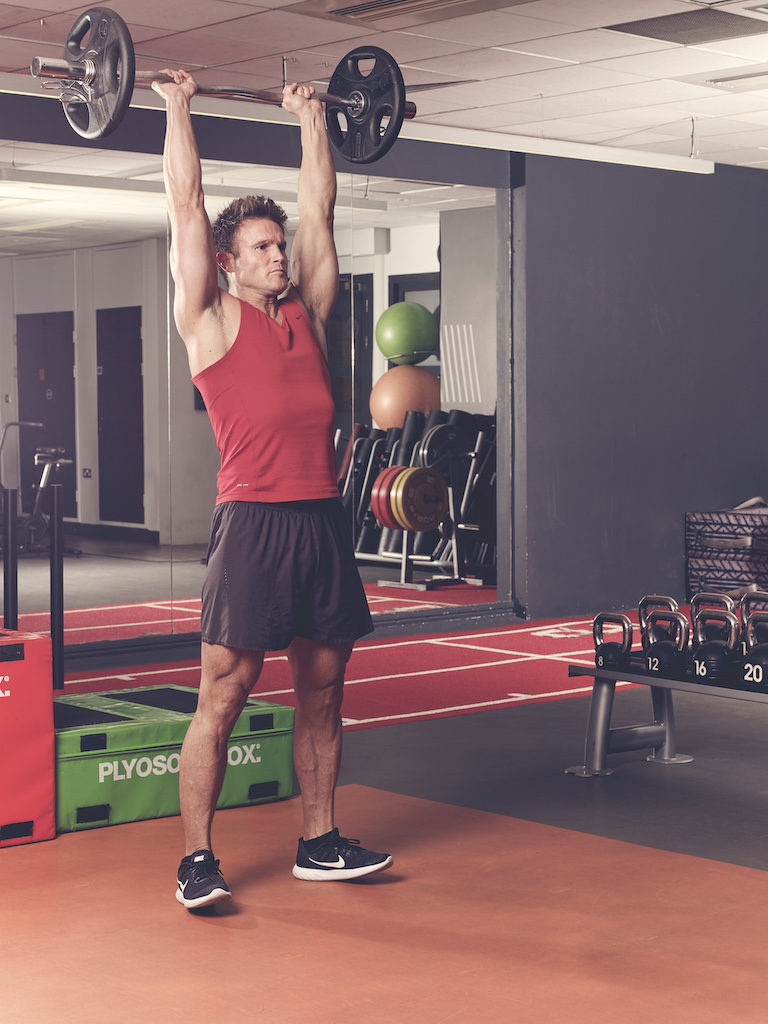 man in red vest and black shorts performing overhead triceps extension in the gym