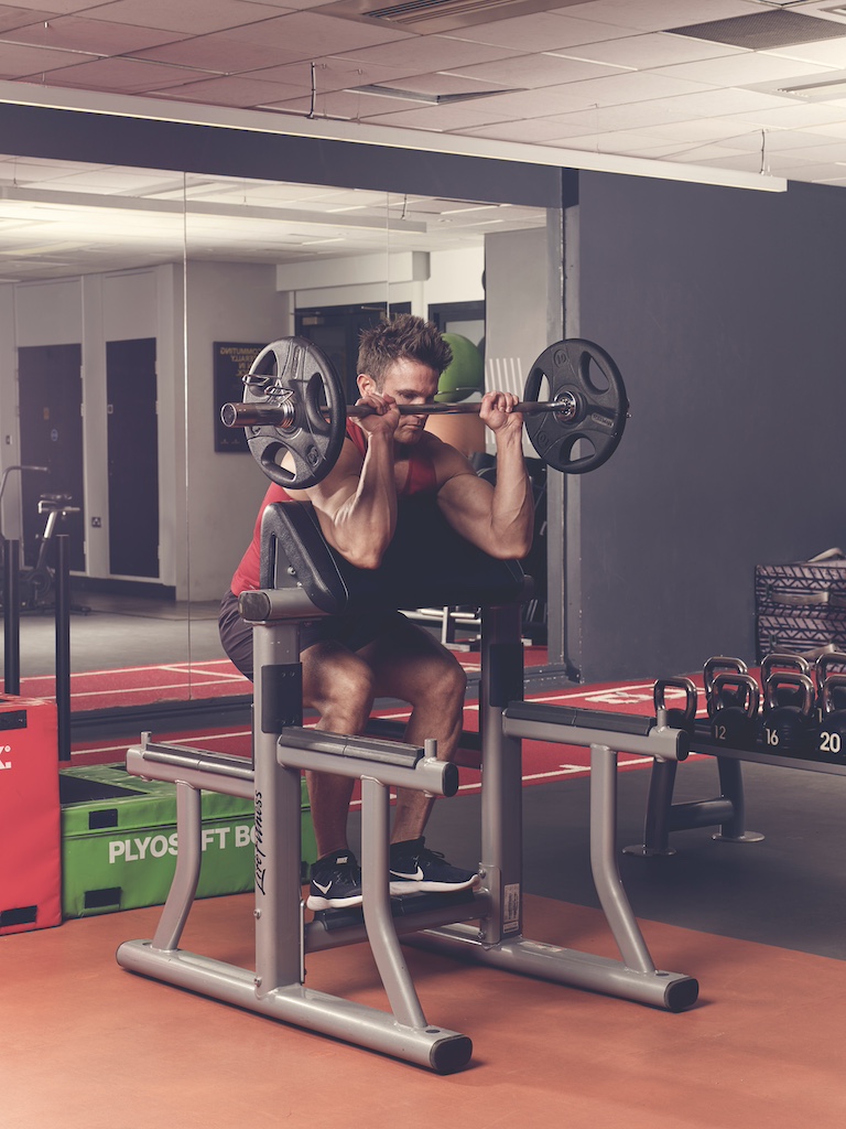 man in red vest and black shorts performing EZ-bar preacher reverse curl in the gym – one of the best barbell arms exercises