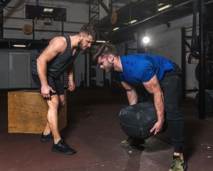 Young muscular sweaty fit instructor or coach man helping veteran man with scars on his skin in rehabilitation process crossfit training and workout in the gym with sandbag and other equipment
