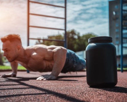 topless man performing a press-up outdoors, with a tub of whey protein in the foreground