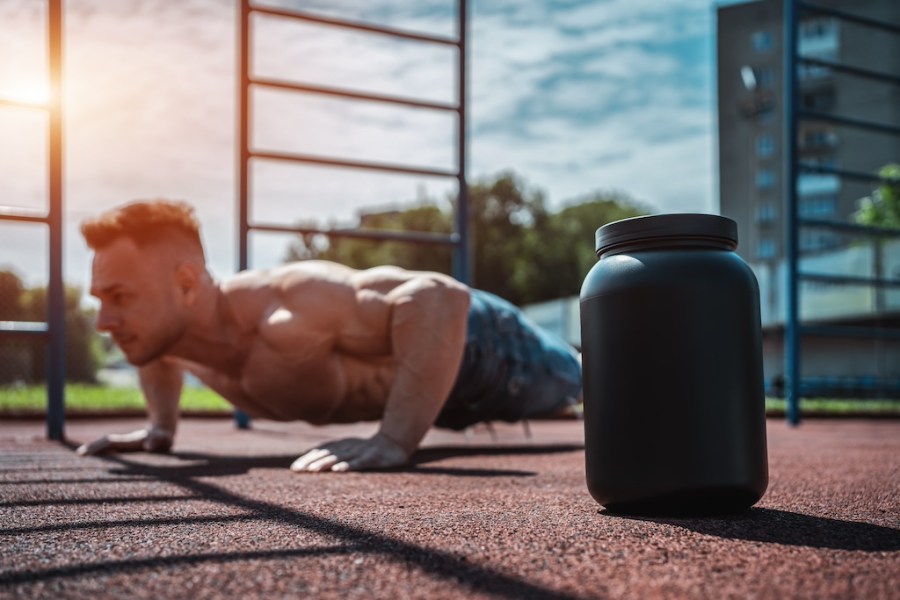 topless man performing a press-up outdoors, with a tub of whey protein in the foreground